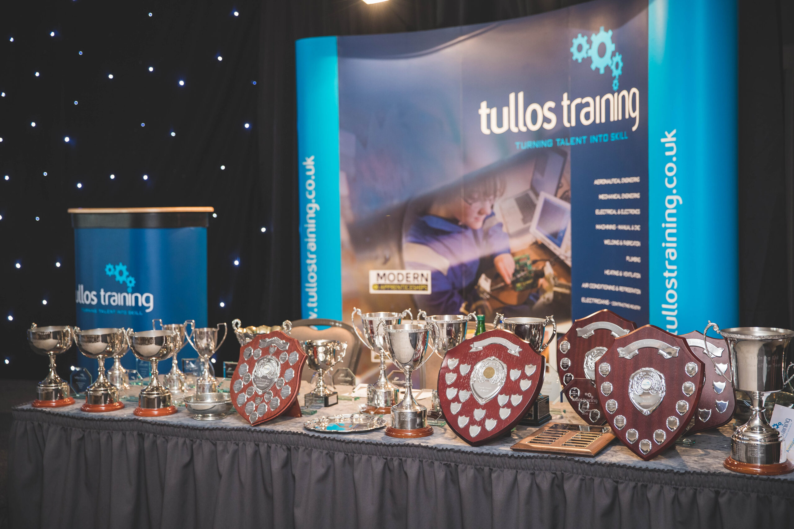 Image shows Tullos Training's Annual Awards displayed on a table with a black tablecloth in front of their branded banner and lectern with a black star cloth backdrop.